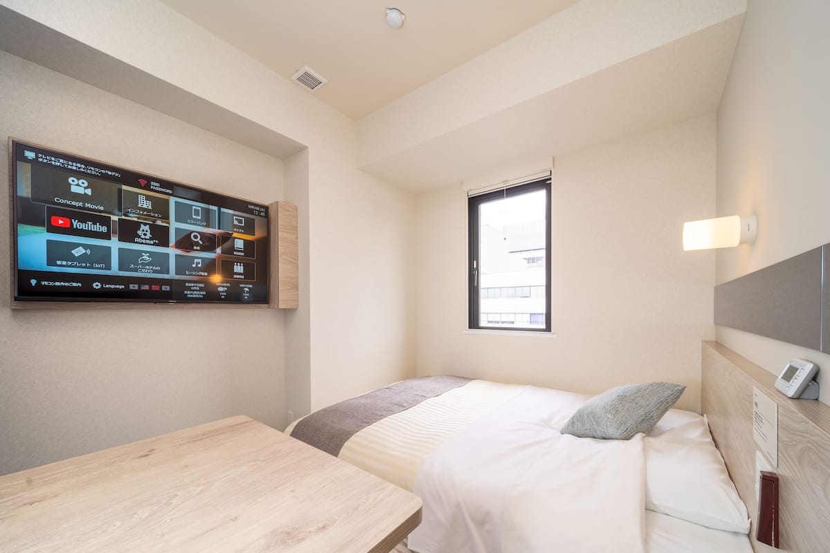 A hotel room with a bed, a bedside table, a TV mounted on the wall displaying various apps, a window with a view of the outside, and a bedside lamp.