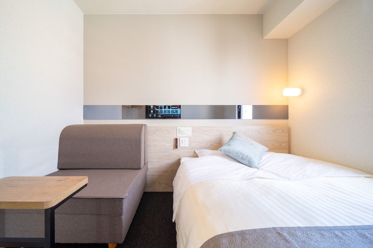 A hotel room with a bed, a sofa, a small table, a wall-mounted control panel, and a light fixture.