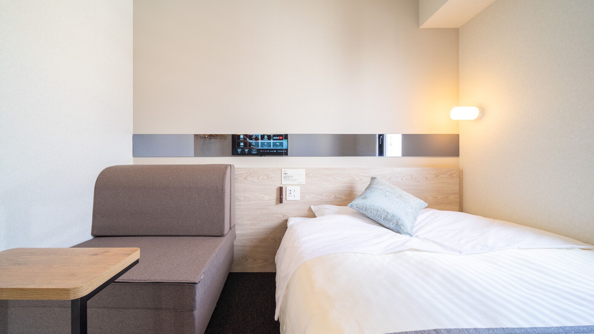 A hotel room with a bed, a sofa, a small table, and a wall-mounted control panel.