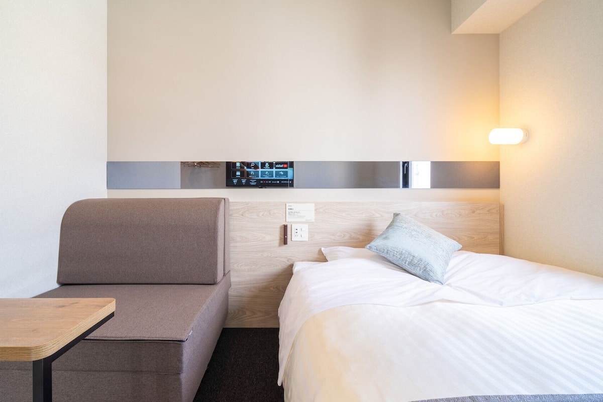 A hotel room with a bed, a sofa, a small table, and a wall-mounted control panel.