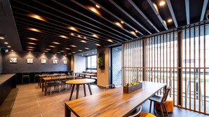 A modern dining area with wooden tables and chairs, a long counter, and large windows offering a view of the outside.