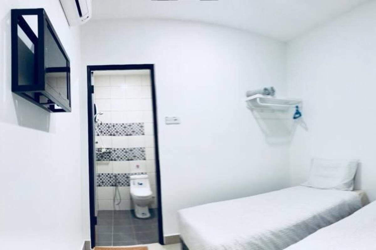 Una stanza piccola e pulita con due letti, una sedia, un televisore, uno specchio e un bagno visibile attraverso una porta aperta.