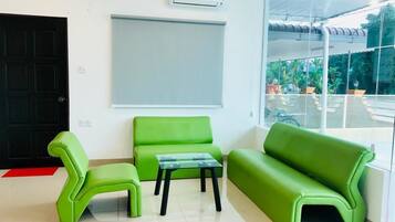 Un área de recepción con una puerta negra, un sofá verde, un sillón verde y una mesa de centro de vidrio. Hay una pizarra en la pared y un equipo de aire acondicionado encima.