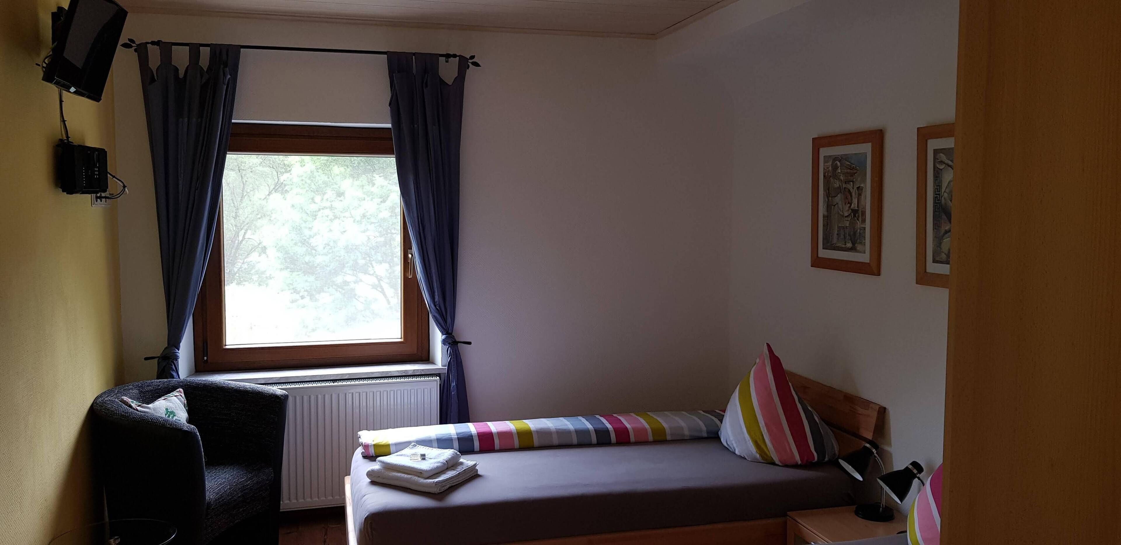 A bedroom with a bed, a chair, a window with curtains, a radiator, and framed pictures on the wall.