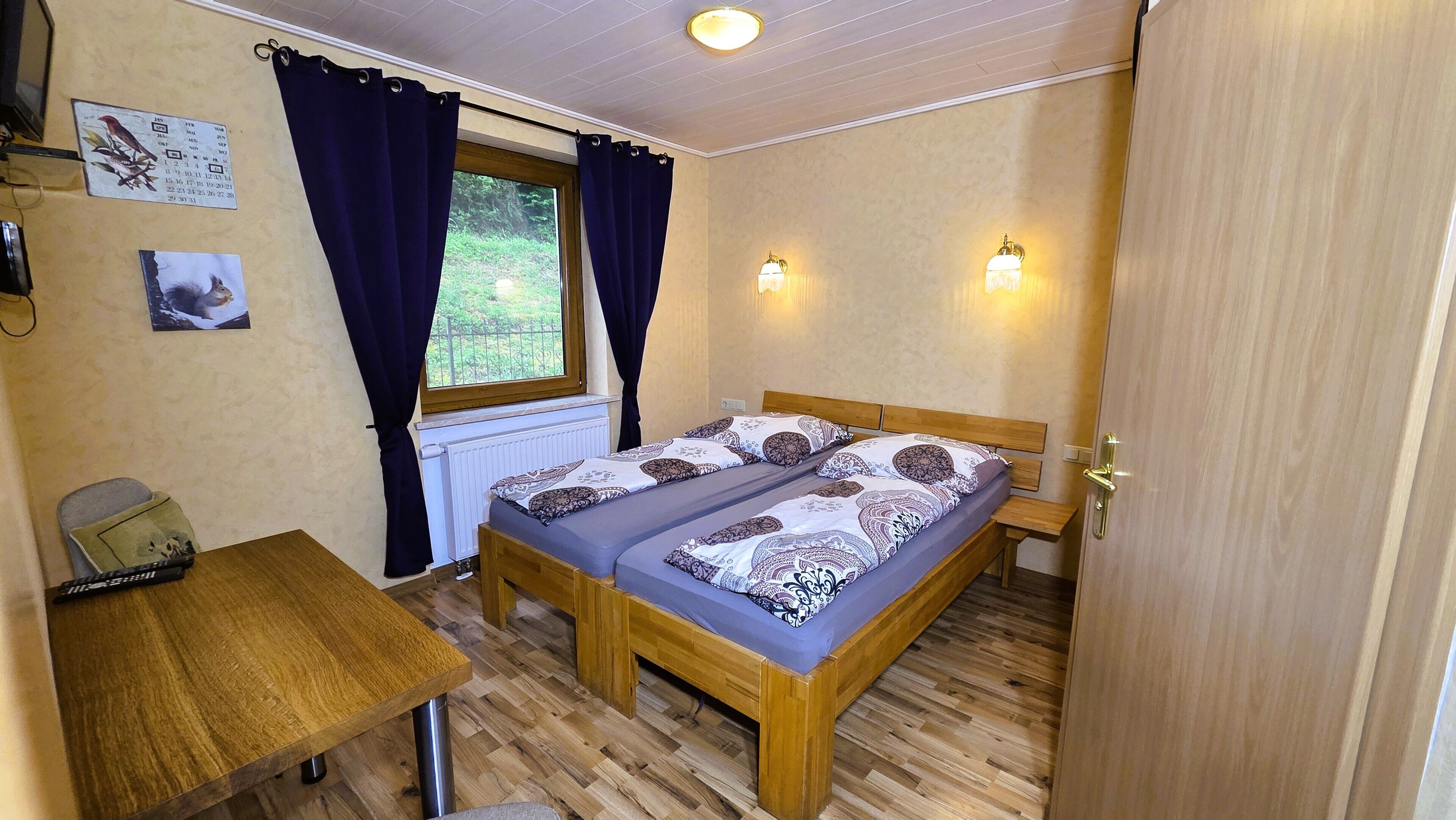 A bedroom with a double bed, a small table, a chair, a window with curtains, and wall-mounted lights.
