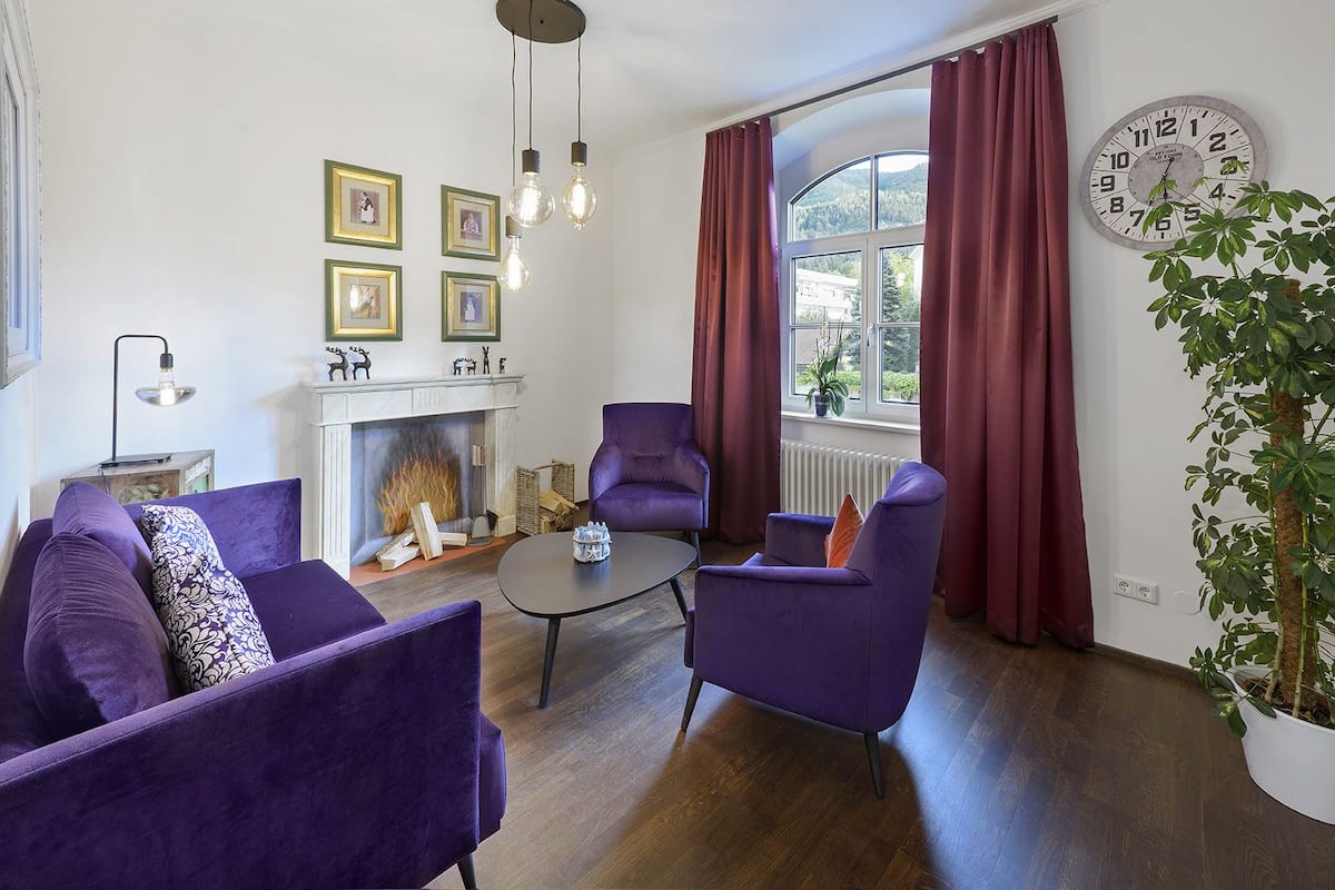 Un salon avec des meubles violets, une cheminée et une grande fenêtre avec des rideaux.