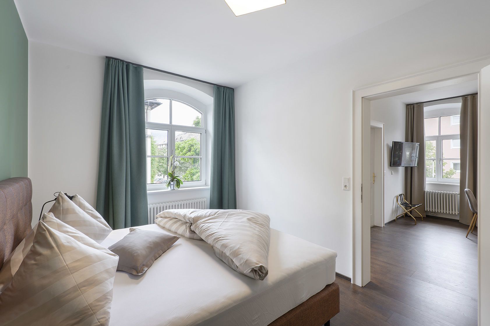 Un dormitorio con cama, ventana con cortinas, radiador, televisor montado en la pared y puerta que da a otra habitación.