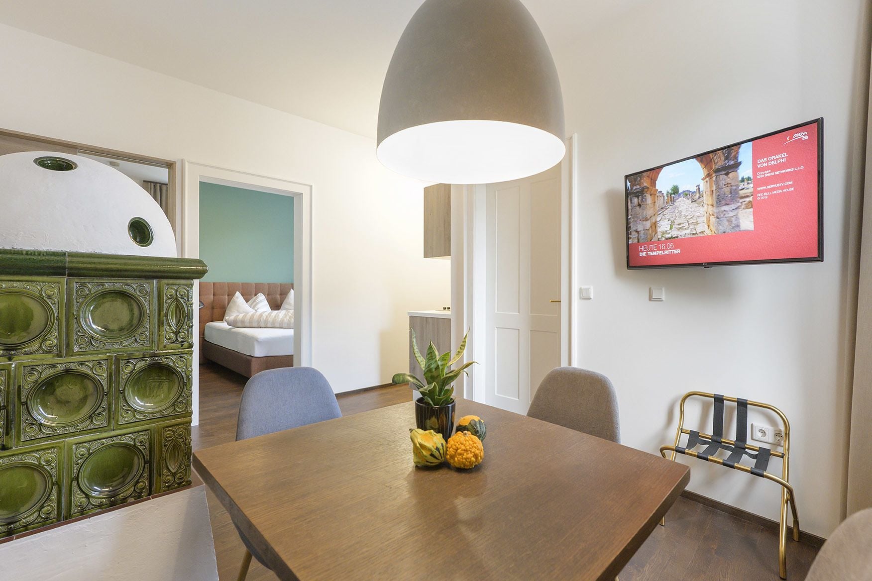 A hotel room with a wooden dining table, a green decorative cabinet, a bed with white pillows, a grey sofa, a TV on the wall, and a plant on the table.