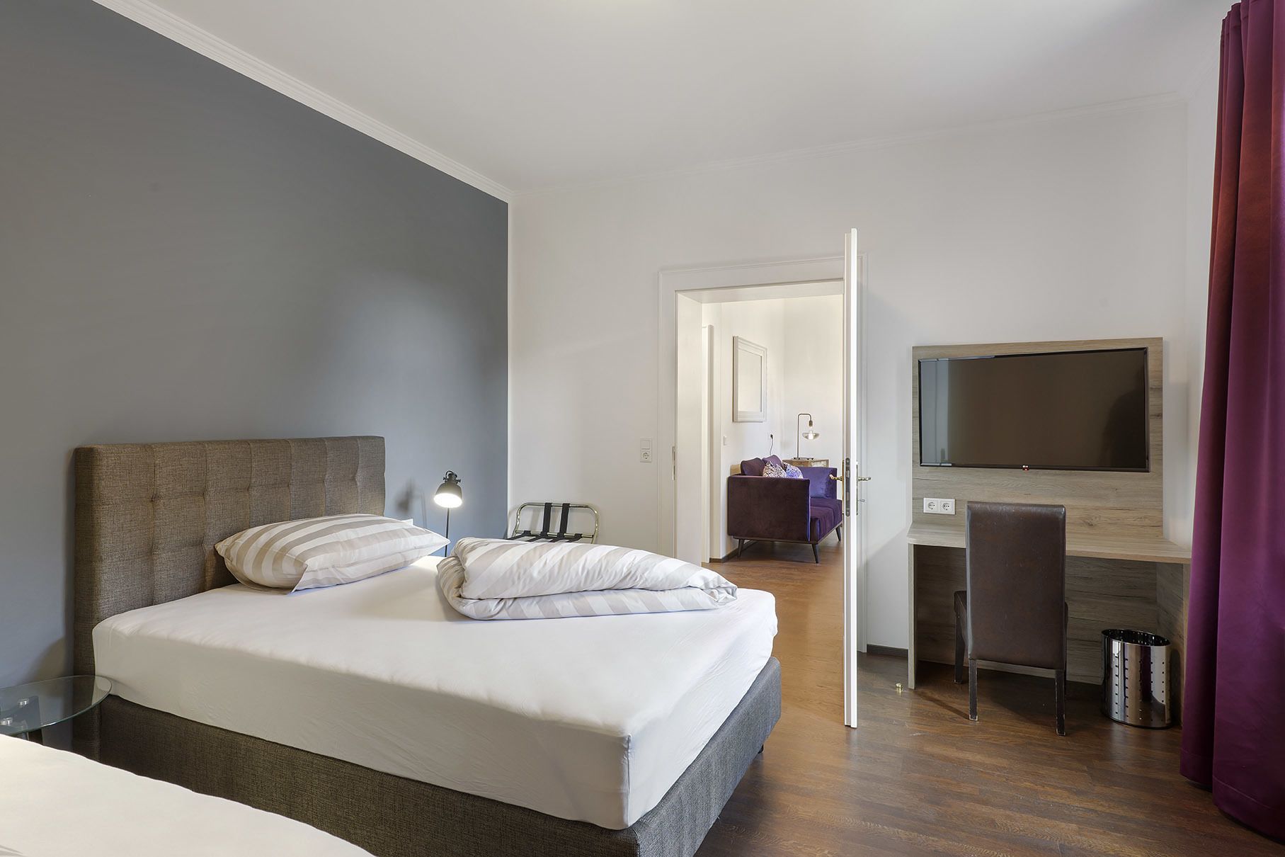 A hotel room with a bed, a desk, a chair, a TV, and a trash can.