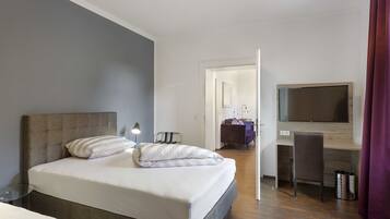 A hotel room with a bed, a desk, a chair, a TV, and a trash can.