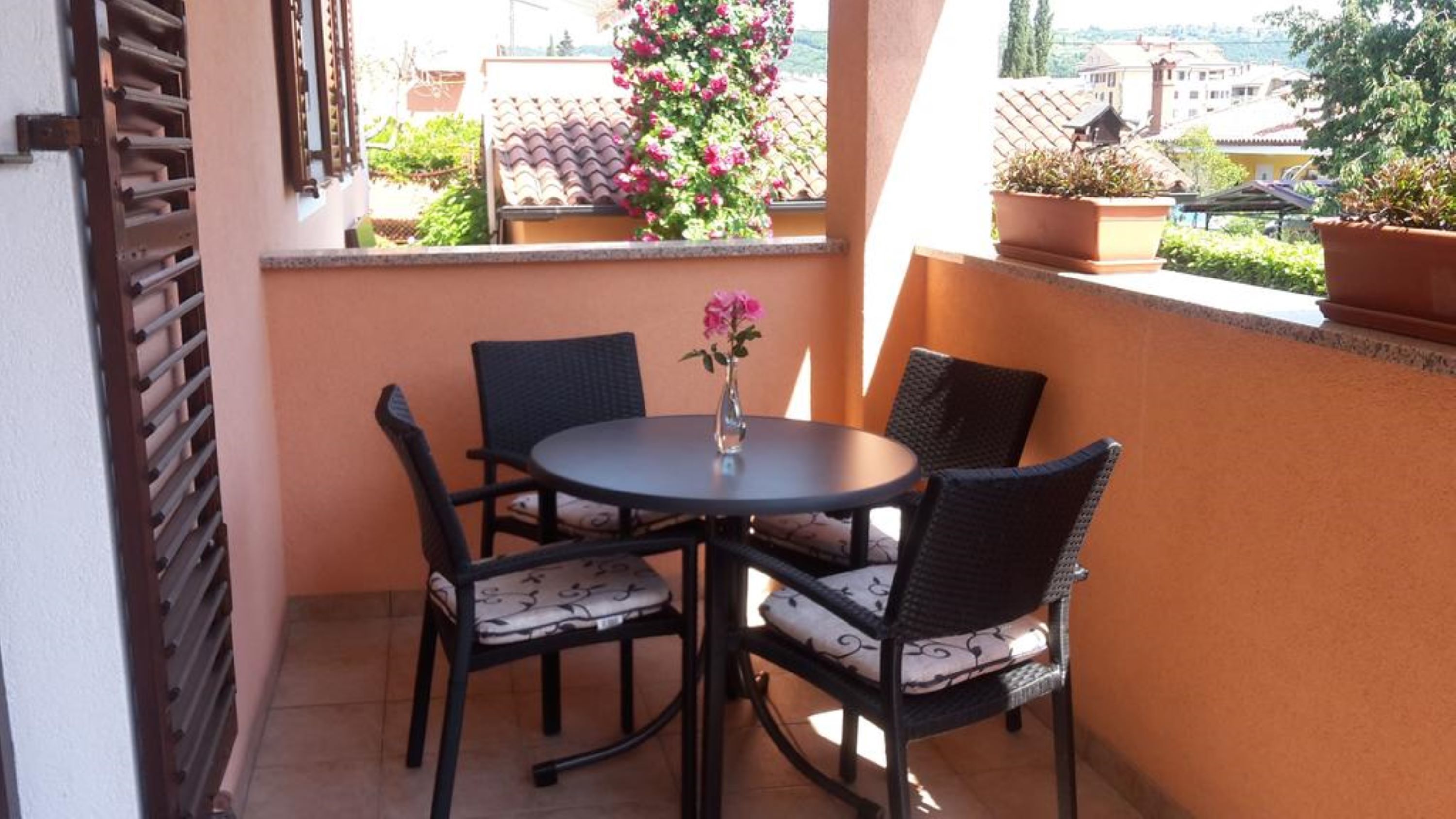A small balcony with a round table and four chairs, a potted plant, and a view of buildings in the background.