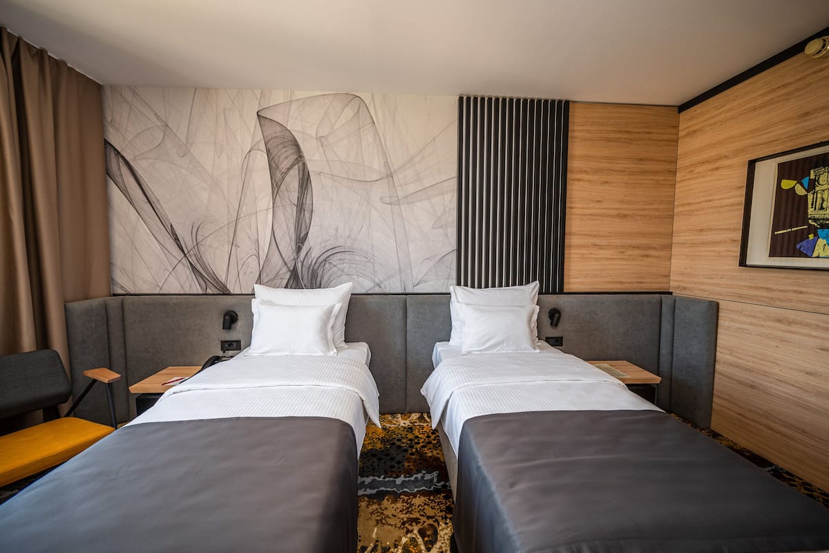A hotel room with two beds, a large abstract painting on the wall, a wooden accent wall, and a small table.