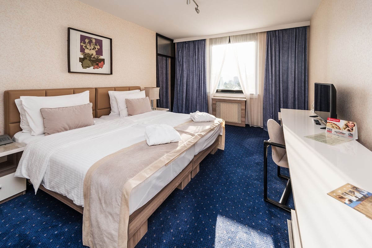 A hotel room with a large bed, a desk with a computer, a window with curtains, and a framed picture on the wall.