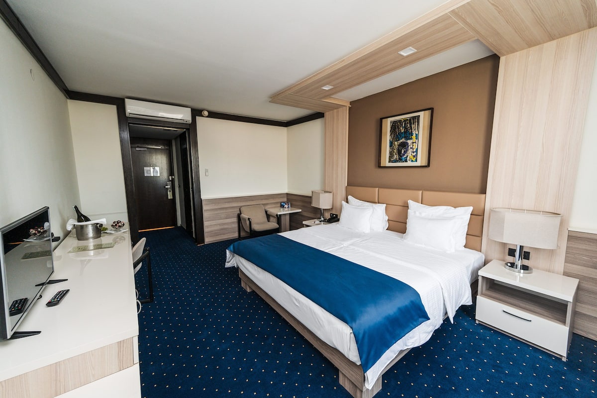 A hotel room with a bed, desk, chair, and a painting on the wall.