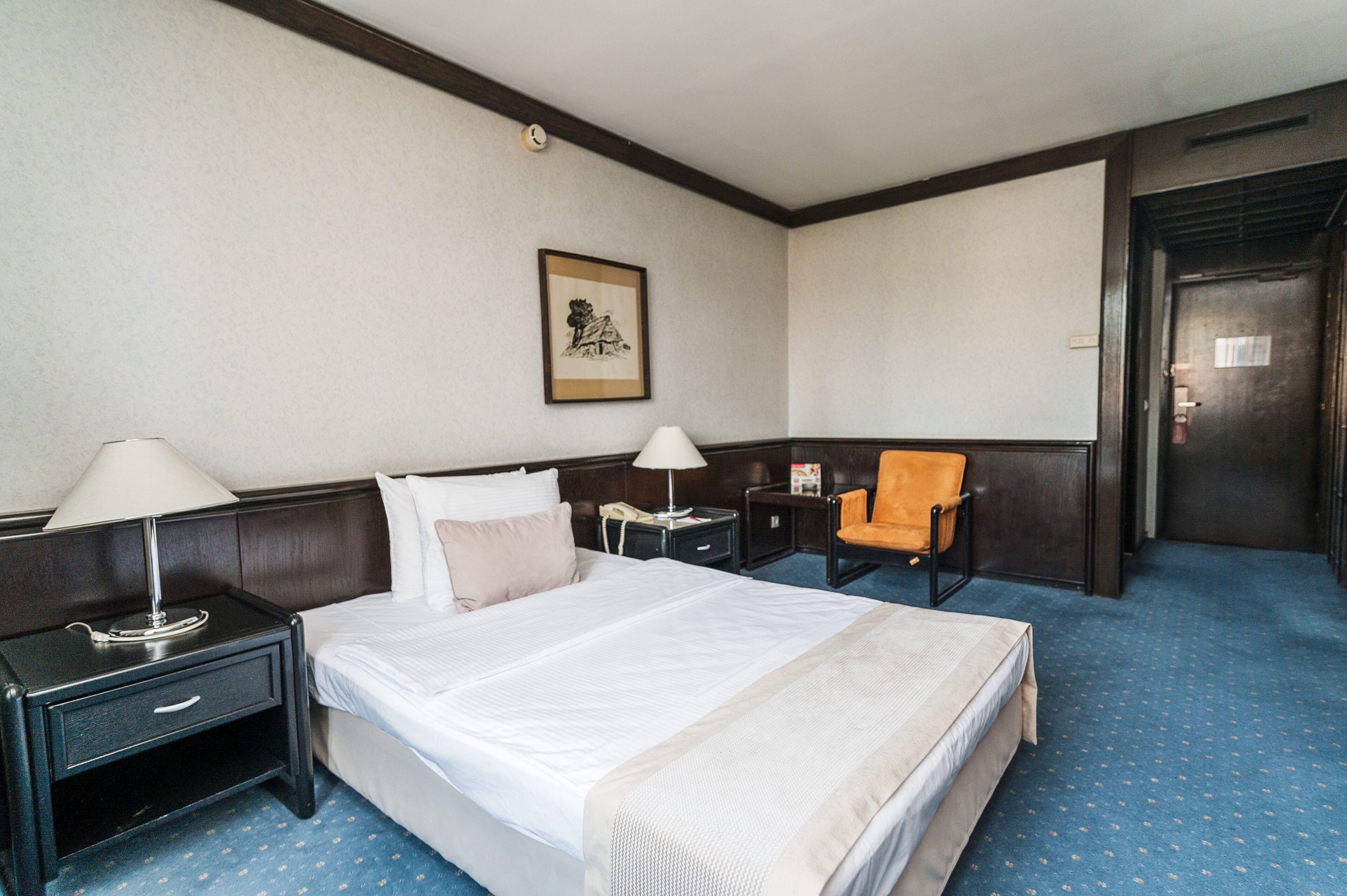 Ein Hotelzimmer mit einem großen Bett, zwei Nachttischen, einem Sessel, einem gerahmten Bild an der Wand und einer Tür.