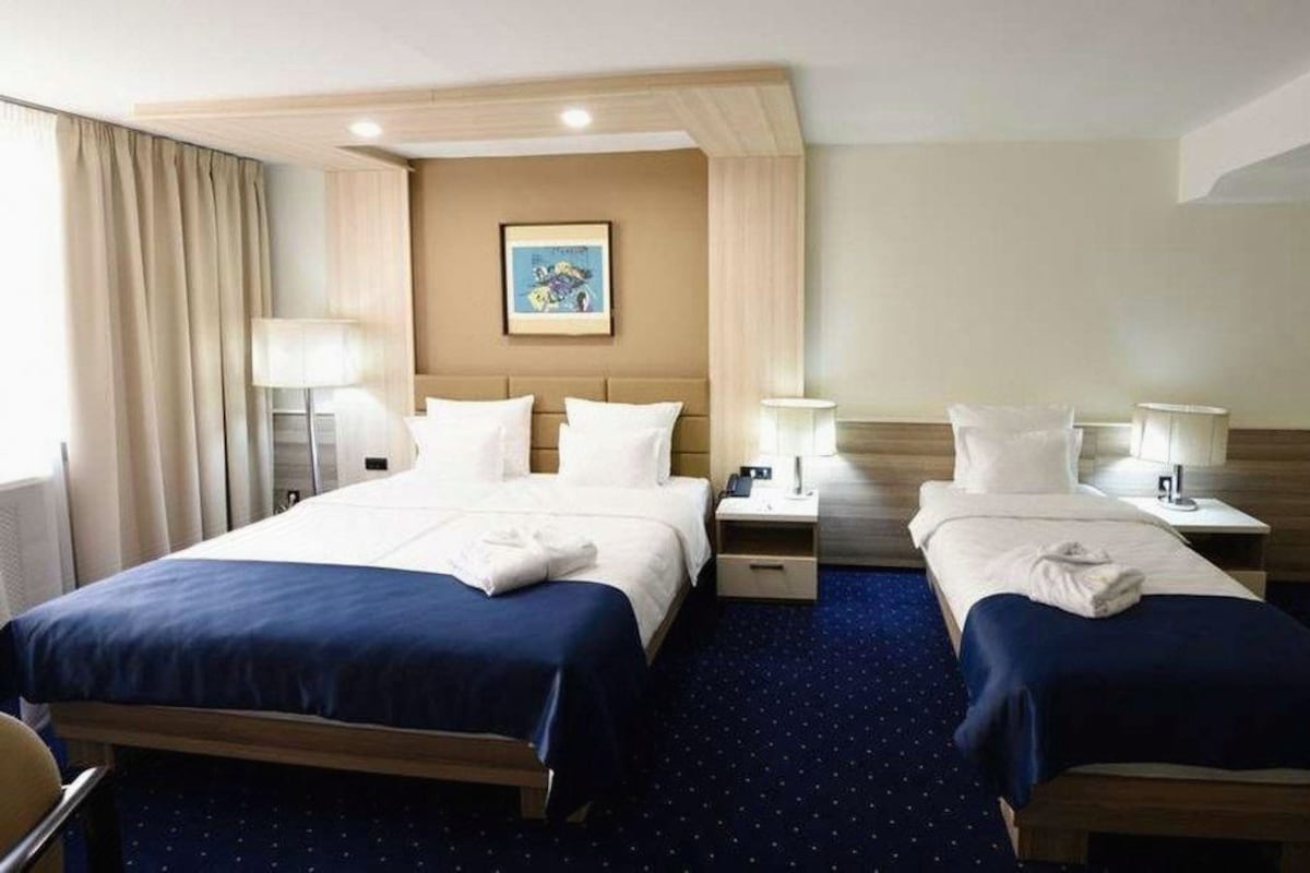 A hotel room with two beds, a large window with curtains, a painting on the wall, and a blue carpet.