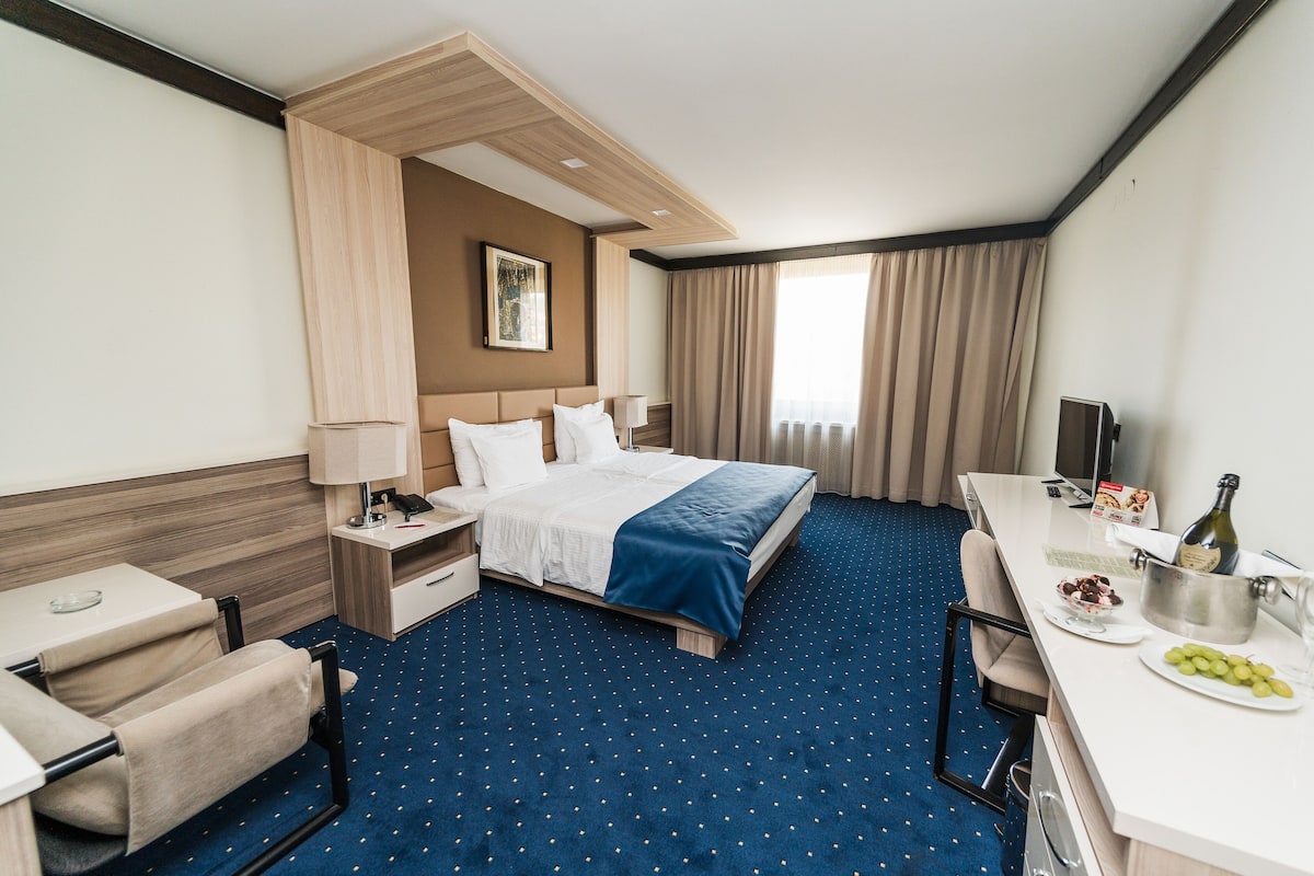 A hotel room with a bed, a desk with a chair, a TV, and a minibar.