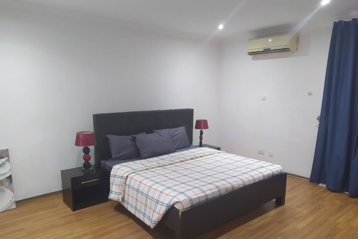 A bedroom with a bed, two bedside tables with lamps, an air conditioner, and a blue curtain.