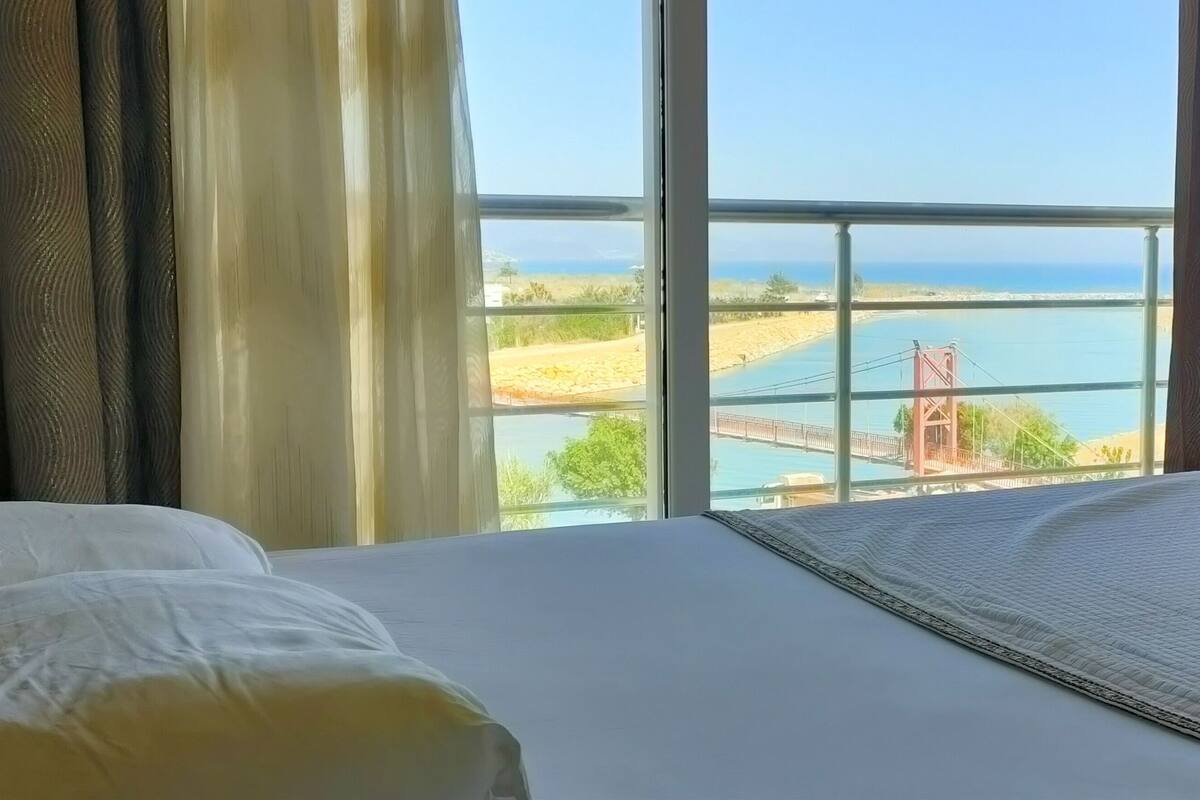 A hotel room with a large window offering a view of a beach and a bridge.