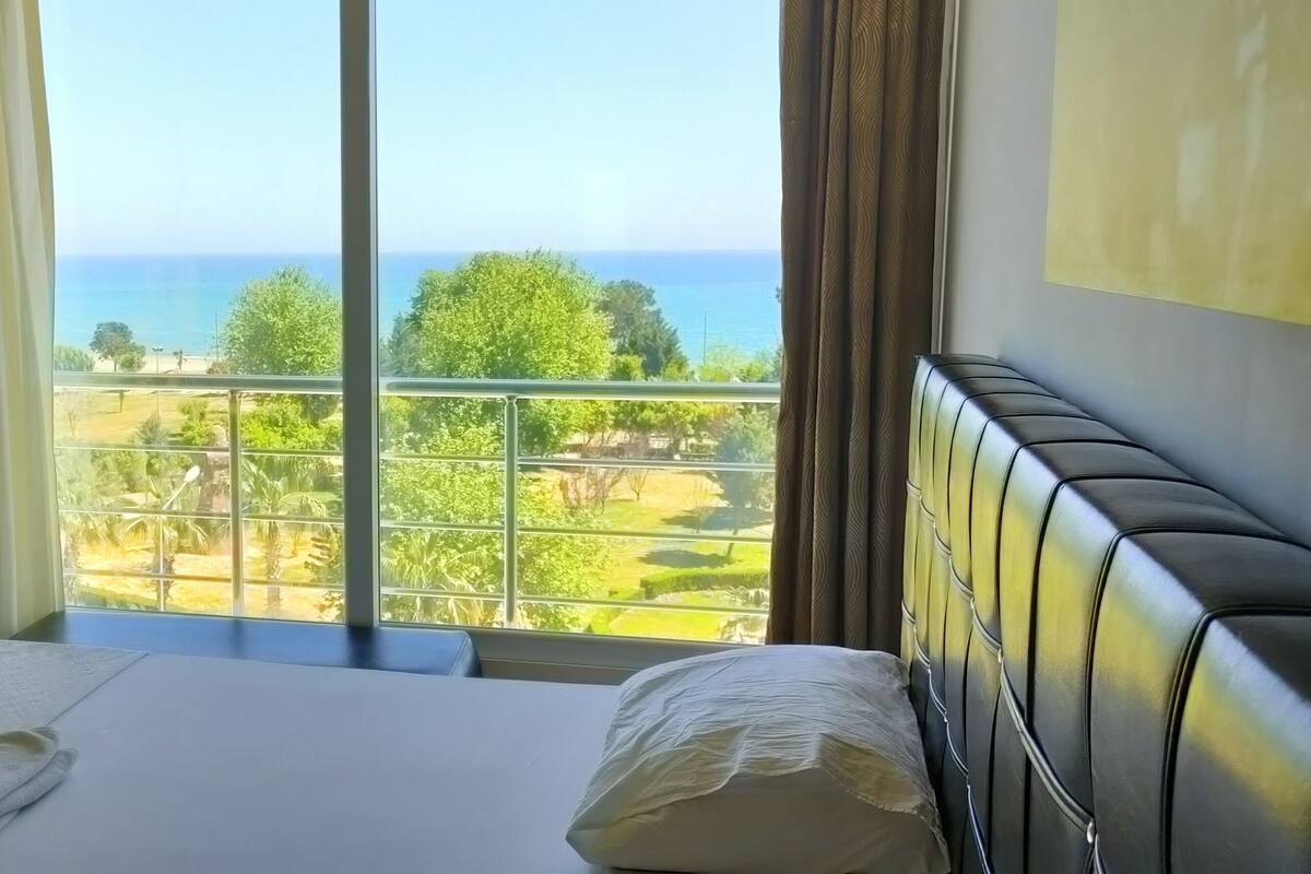 A hotel room with a large window offering a view of the sea and greenery.