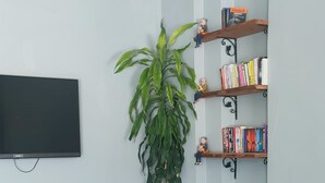 Una sala de estar con dos sofás verdes, una mesa de centro de madera, una planta grande en maceta, una estantería llena de libros y un televisor de pantalla plana montado en la pared.