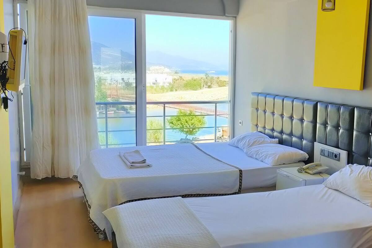 A hotel room with two beds, a large window with a view of the beach, a bedside table with a phone, and a yellow accent wall.
