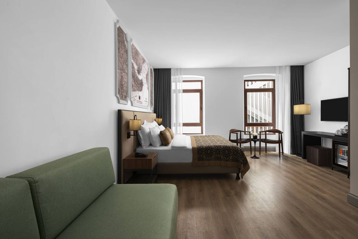 A hotel room with a bed, a green sofa, a desk with a lamp, and a TV.