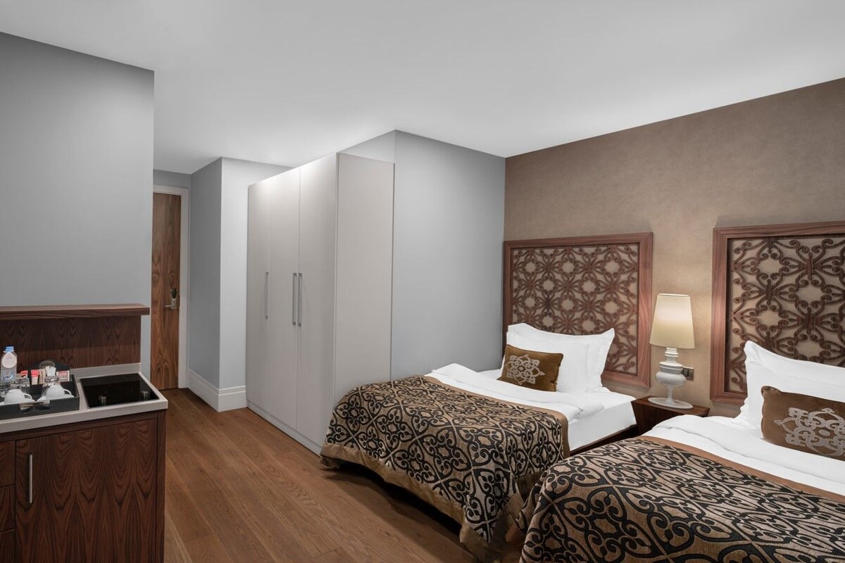 Une chambre d’hôtel avec une kitchenette, deux lits recouverts de couvre-lits à motifs, une tête de lit décorative et une table de chevet avec une lampe.