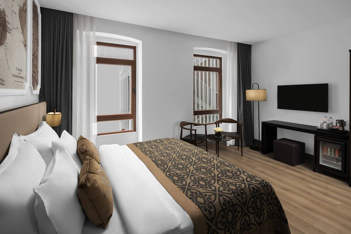 A hotel room with a large bed, a desk, a chair, a TV, and a bookshelf.