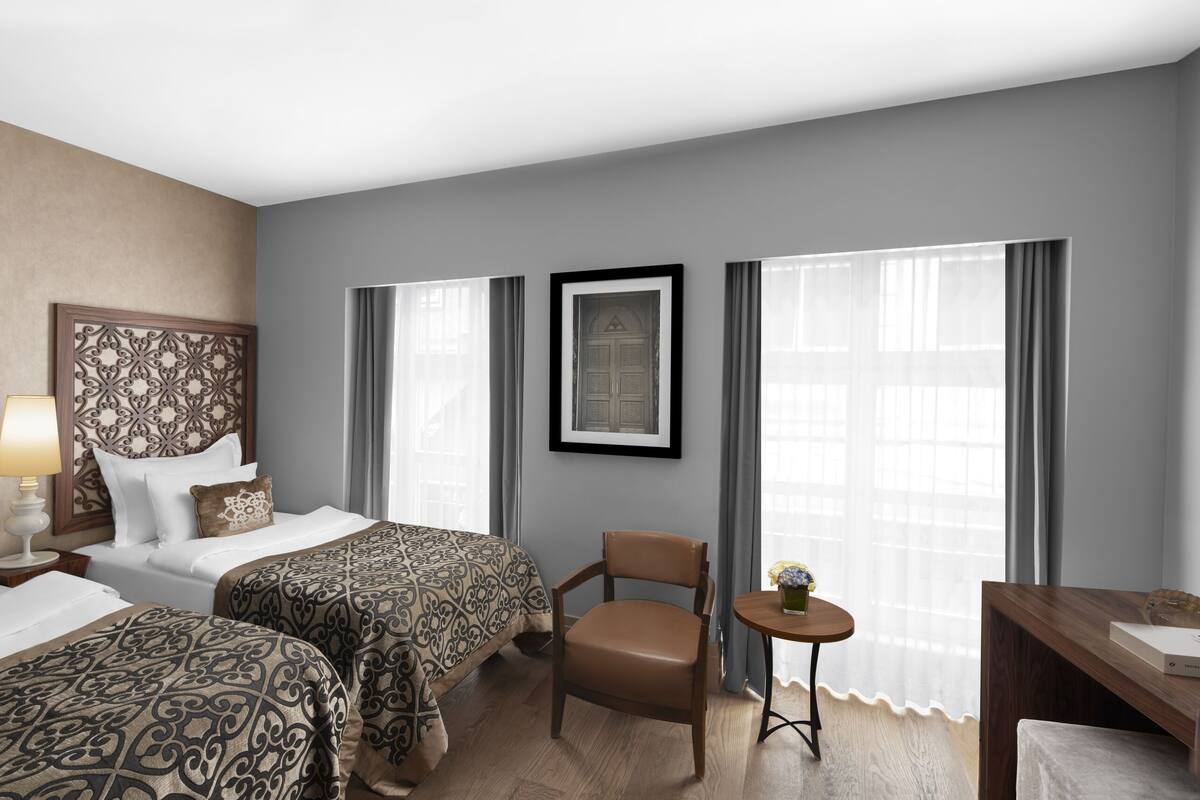 A hotel room with two beds, a desk, a chair, a TV, and large windows with curtains.