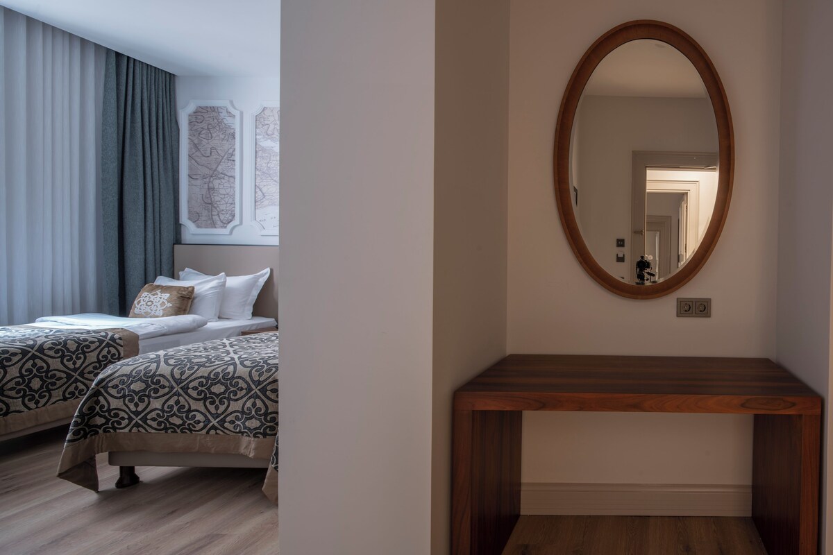 Habitación de hotel con dos camas, una mesita de madera y un espejo grande en la pared.