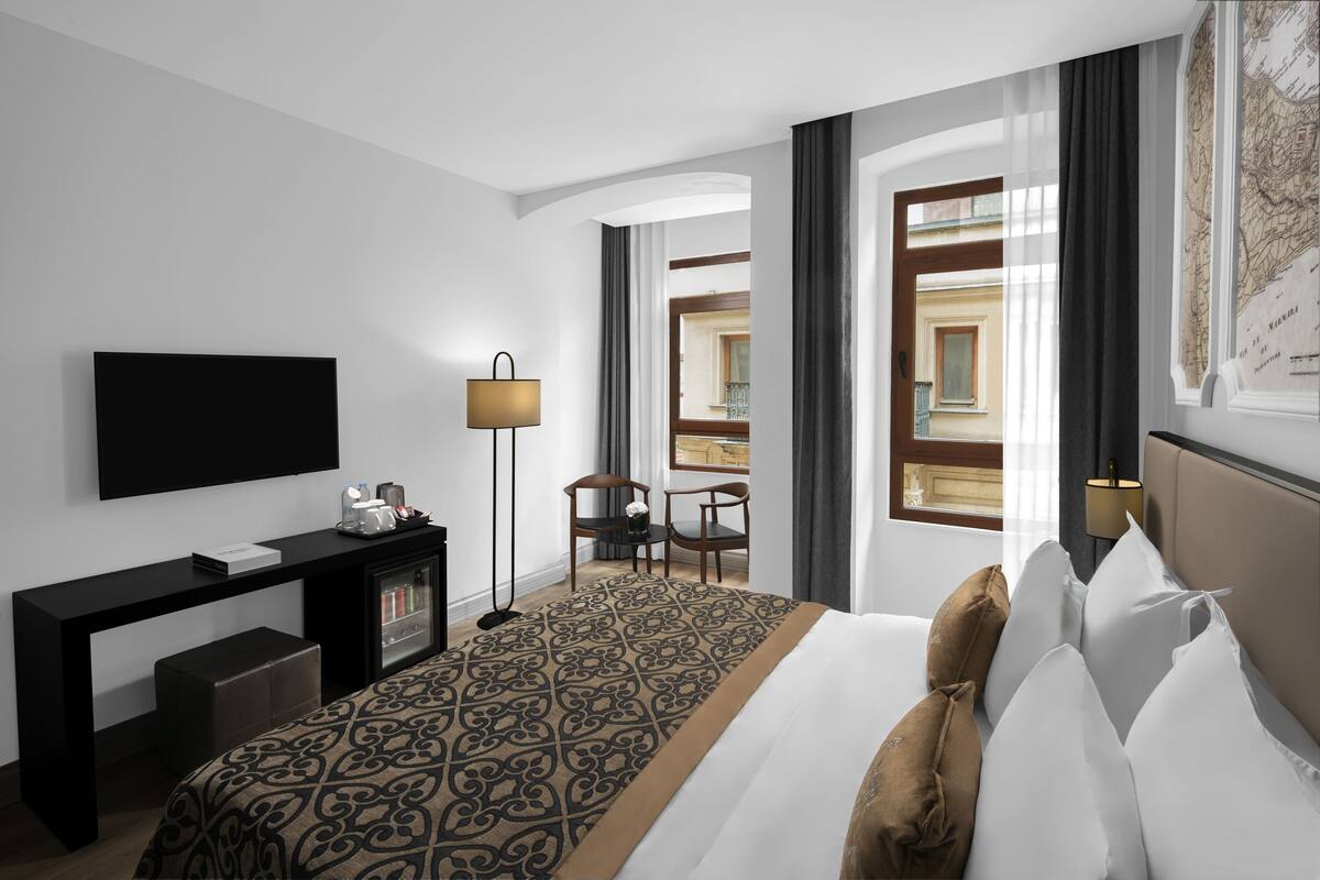 A modern hotel room with a large bed, a flat-screen TV, a desk with a lamp, and a view of the city through large windows.