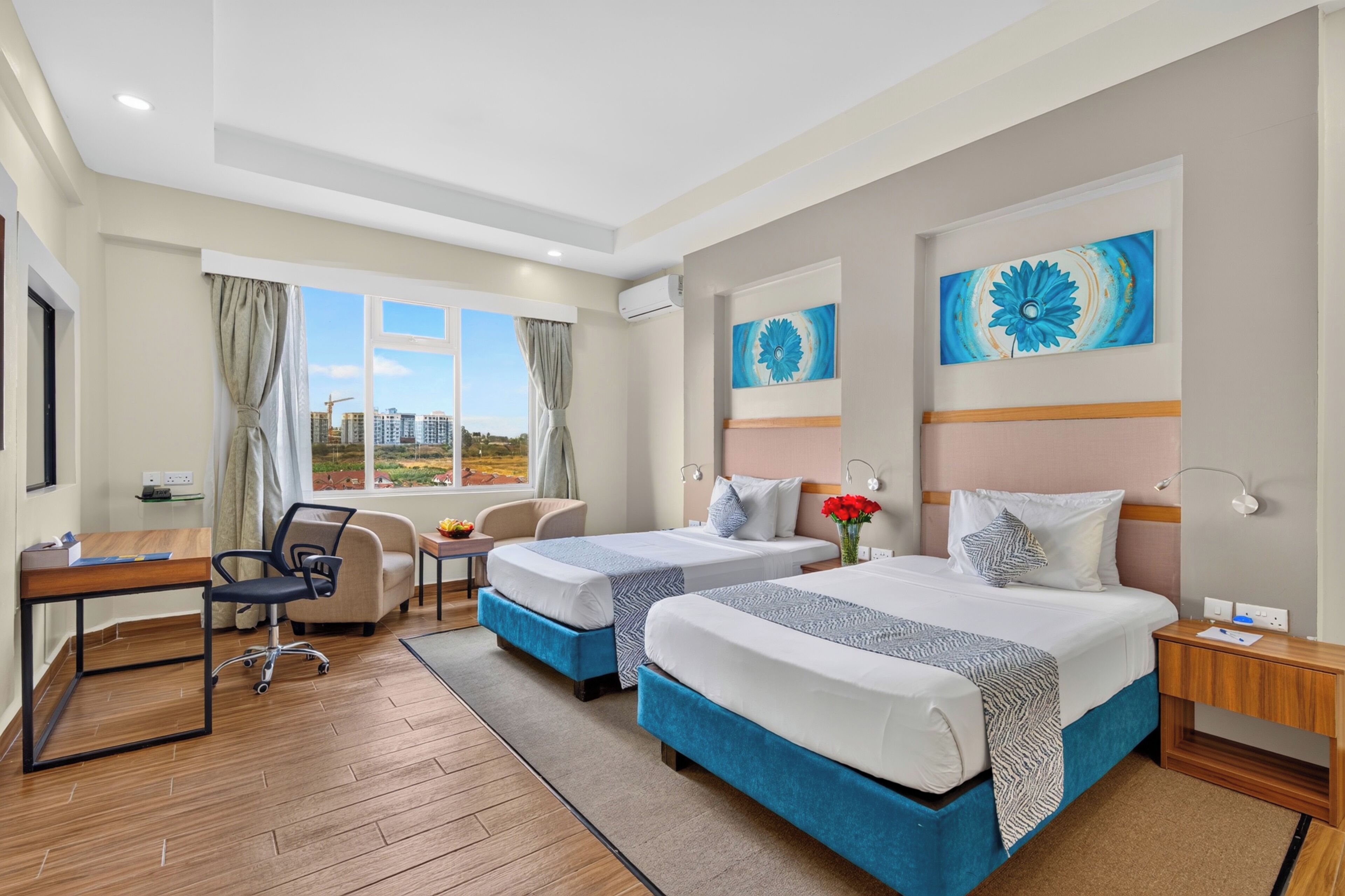 A hotel room with two beds, a desk, a chair, and a view of buildings outside.