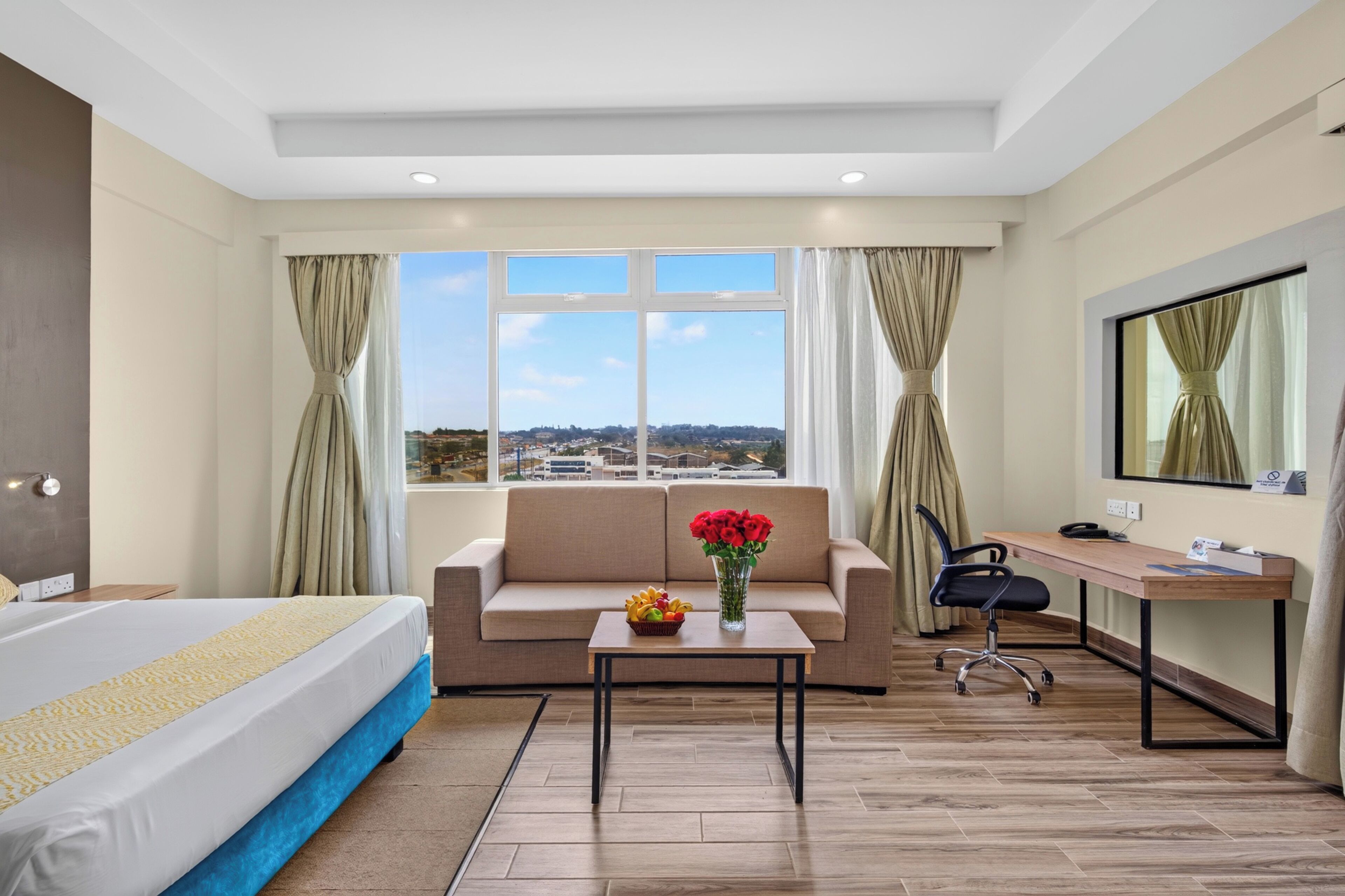 Une chambre d’hôtel comprenant un lit, un canapé, un bureau, une chaise, une petite table avec un vase de fleurs et une grande fenêtre donnant sur un paysage urbain.