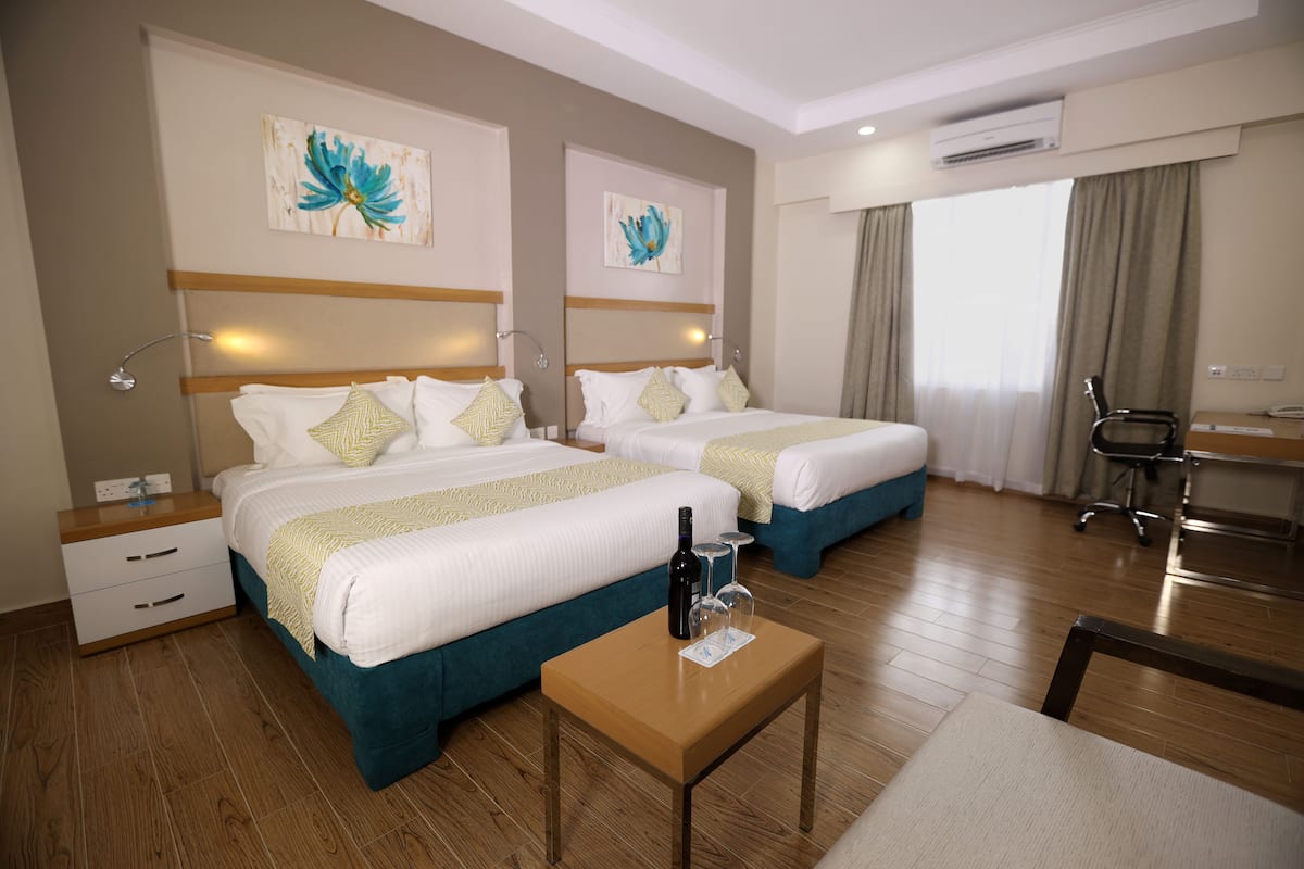 Une chambre d’hôtel avec deux lits, une table de chevet en bois, une petite table avec une bouteille et des verres, et des œuvres d’art accrochées aux murs.