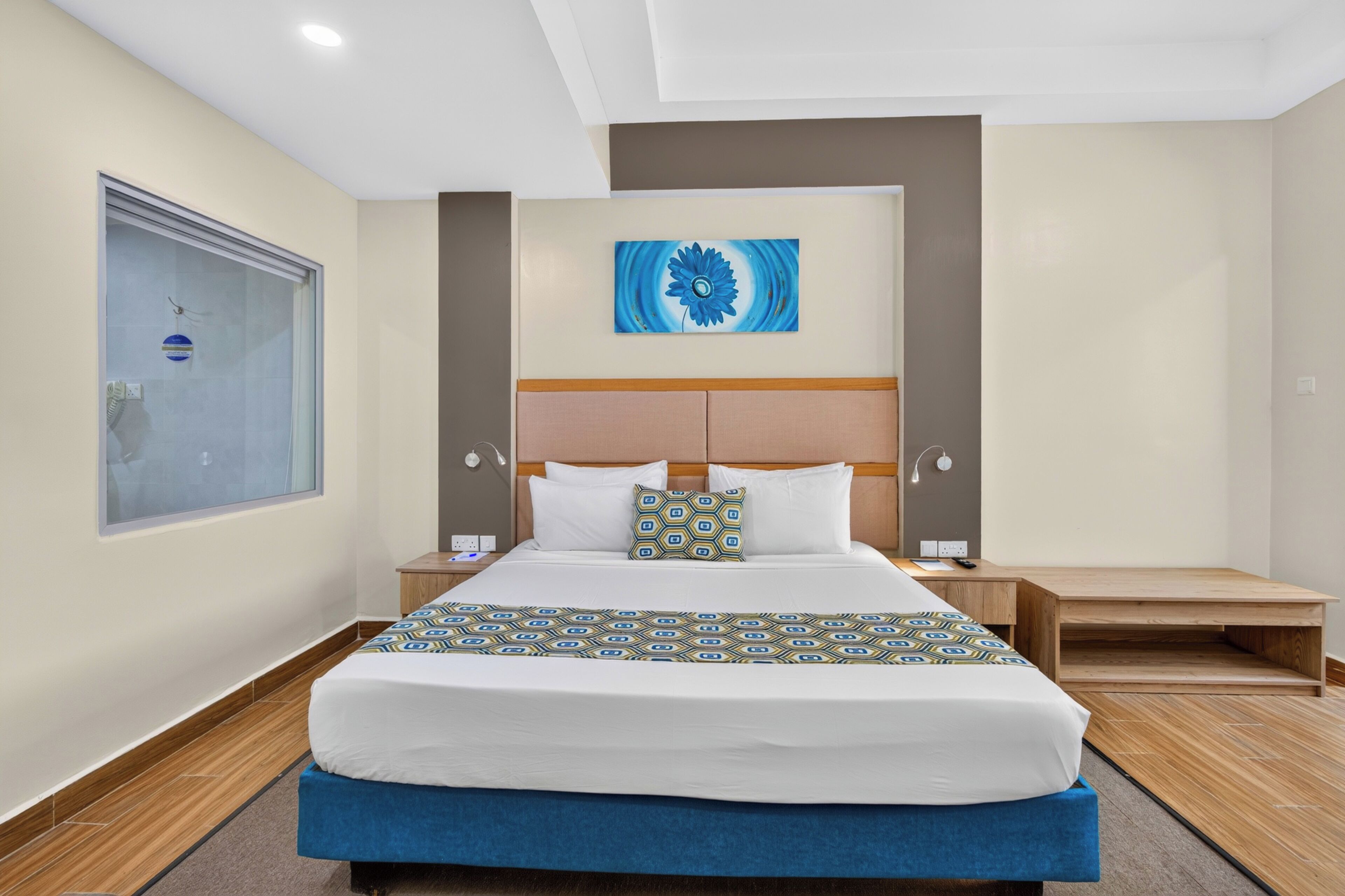 Habitación de hotel con una cama grande, cabecero, mesita de noche y un cuadro en la pared. Se aprecia una ventana con vistas.