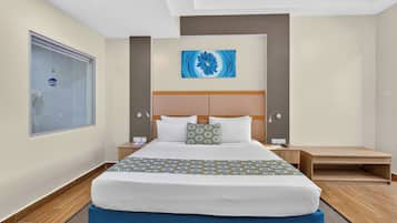 Habitación de hotel con una cama grande, cabecero, mesita de noche y un cuadro en la pared. Se aprecia una ventana con vistas.