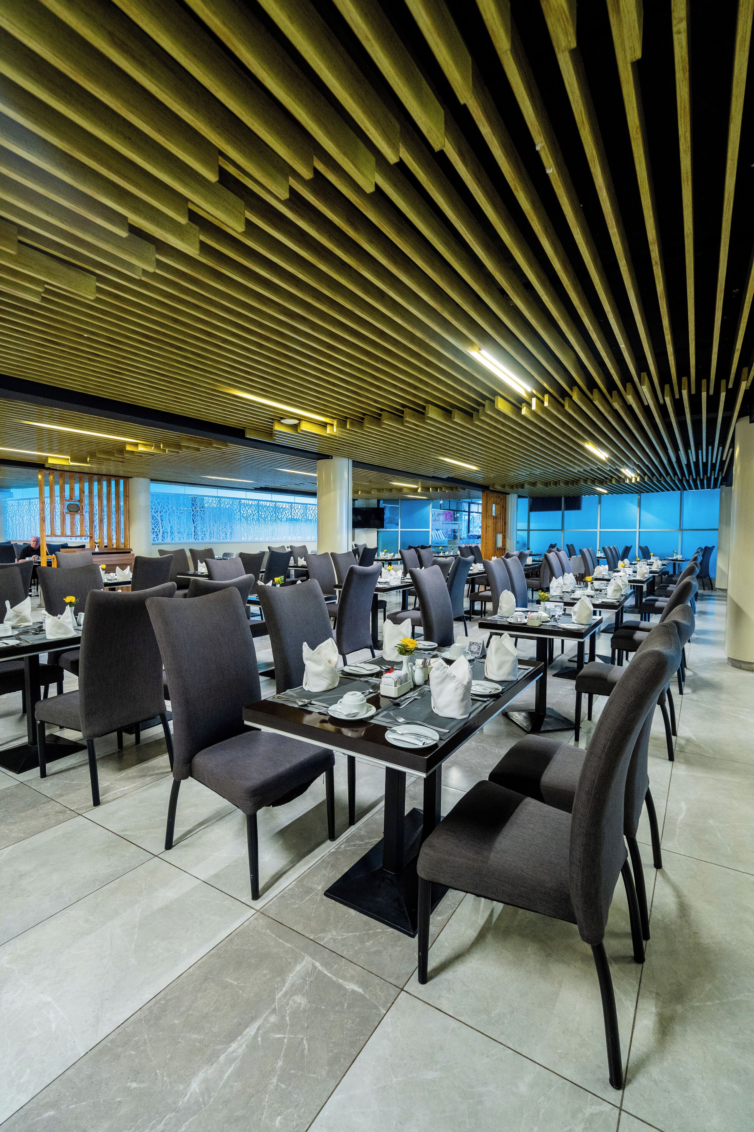 A spacious dining area with modern furniture and a unique ceiling design. Tables are neatly arranged with chairs, and the floor is tiled.