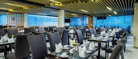 A spacious dining area with modern furniture and a unique ceiling design. Tables are neatly arranged with chairs, and the floor is tiled.