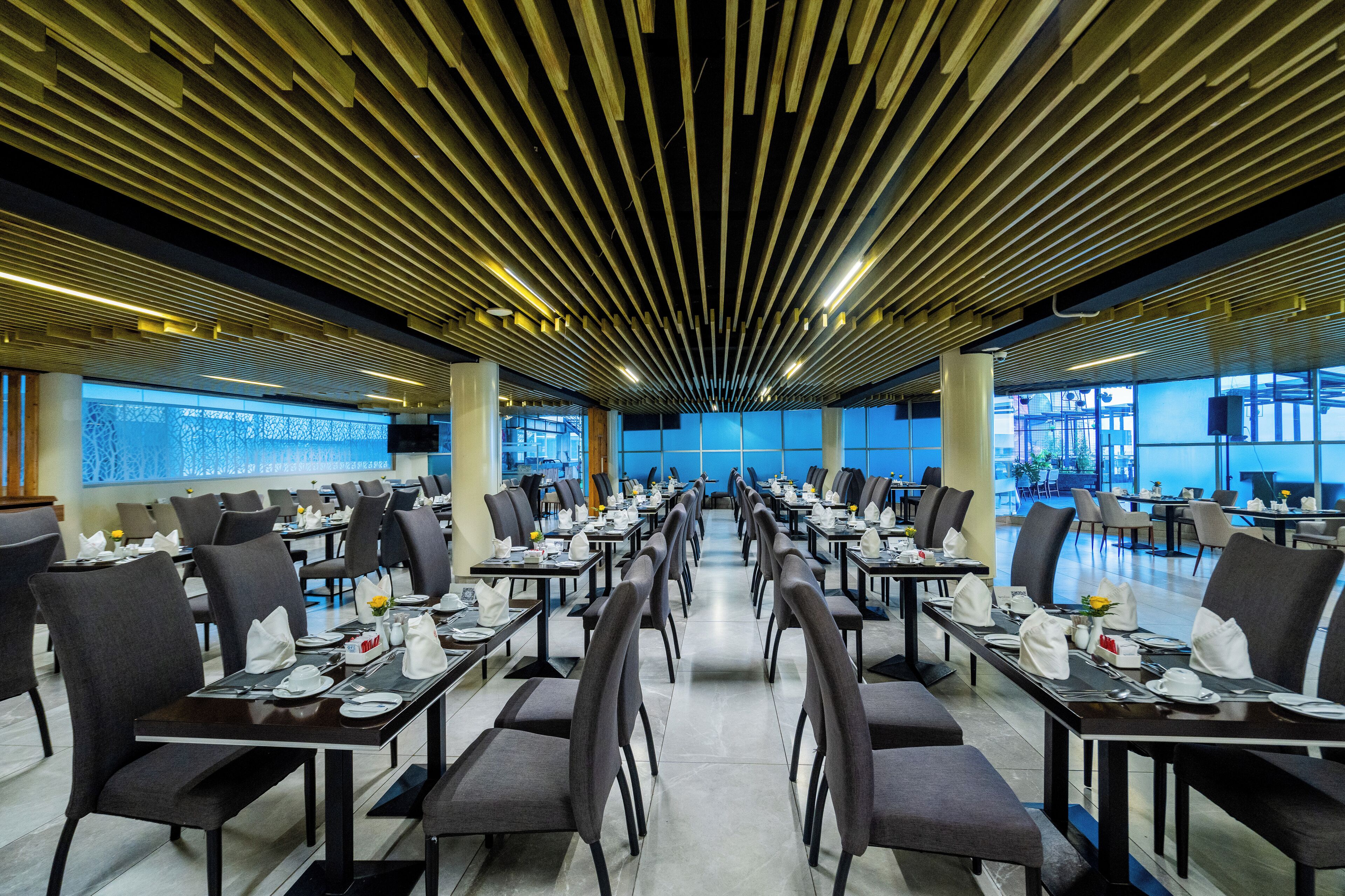 A spacious restaurant with long tables and chairs, a modern ceiling design, and large windows.