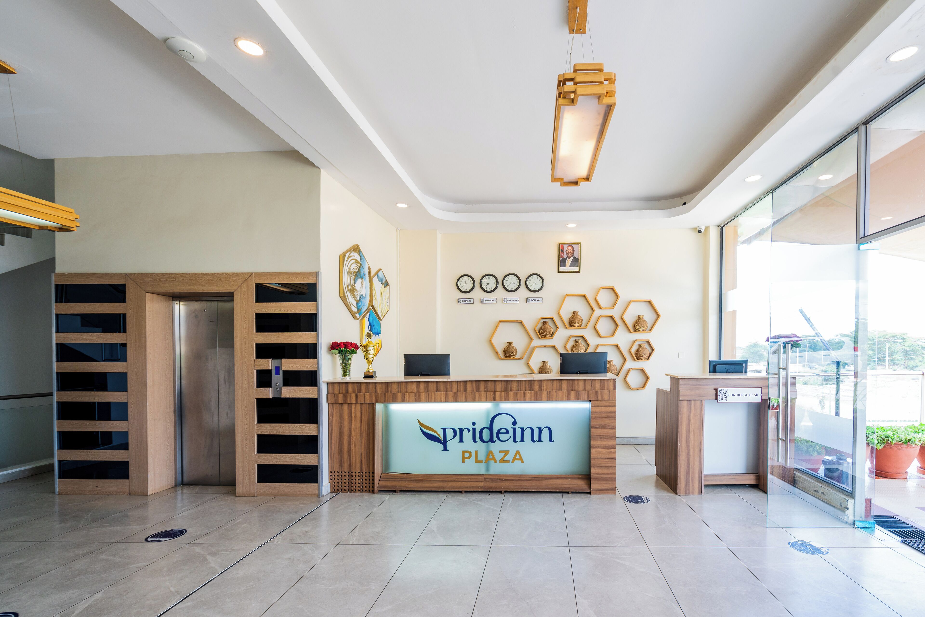 A reception desk with a sign reading "prideinn plaza," a wall with clocks and a framed photo, and a glass door leading outside.