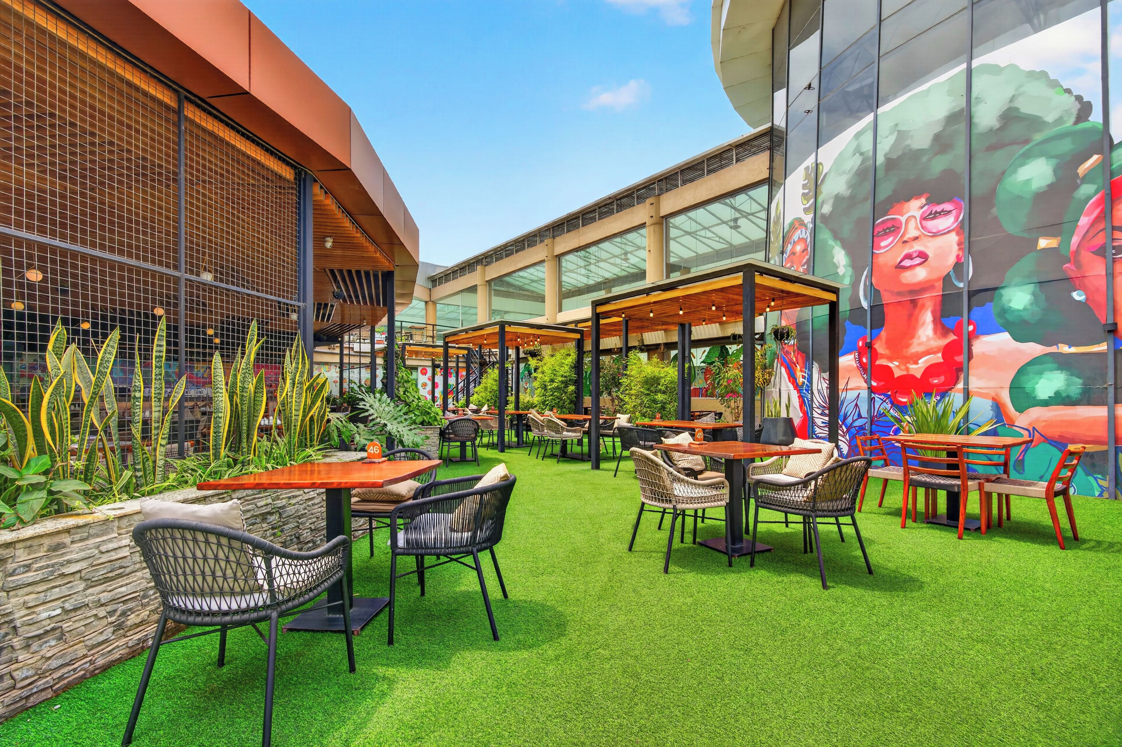 An outdoor seating area with tables and chairs, surrounded by greenery and a mural on the building.