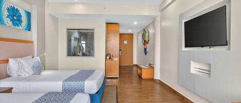 A hotel room with two beds, a television, artwork on the walls, and a hallway leading to a bathroom.
