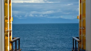 A view through a doorway with a railing, overlooking a calm sea and distant mountains under a cloudy sky.