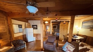 Une pièce chaleureuse avec un plafond en bois, un ventilateur de plafond, un réfrigérateur, un fauteuil à motifs, un canapé agrémenté d’un coussin à fleurs et une table à manger avec des chaises.