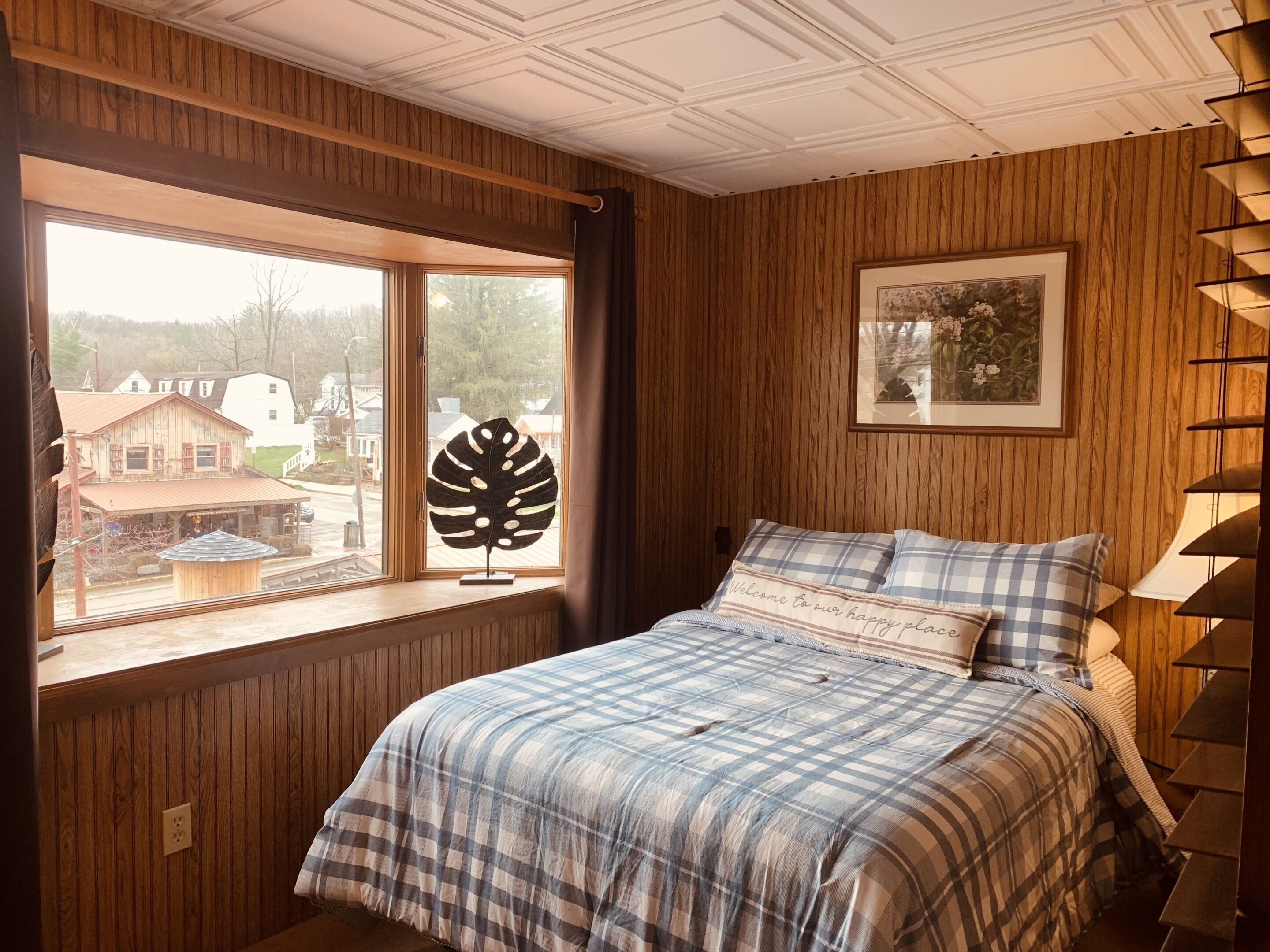 A bedroom with a large window, a bed with plaid bedding, a framed picture on the wall, and a wooden ceiling.