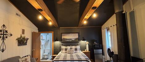 A bedroom with a bed, a sofa, a ceiling fan, a fireplace, and a small kitchen area.