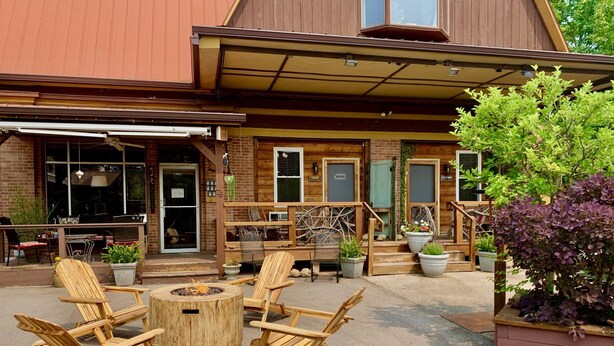 Un espace extérieur aménagé avec des chaises en bois et une table ronde, un bâtiment en briques doté d’une terrasse en bois, et des plantes en pot.