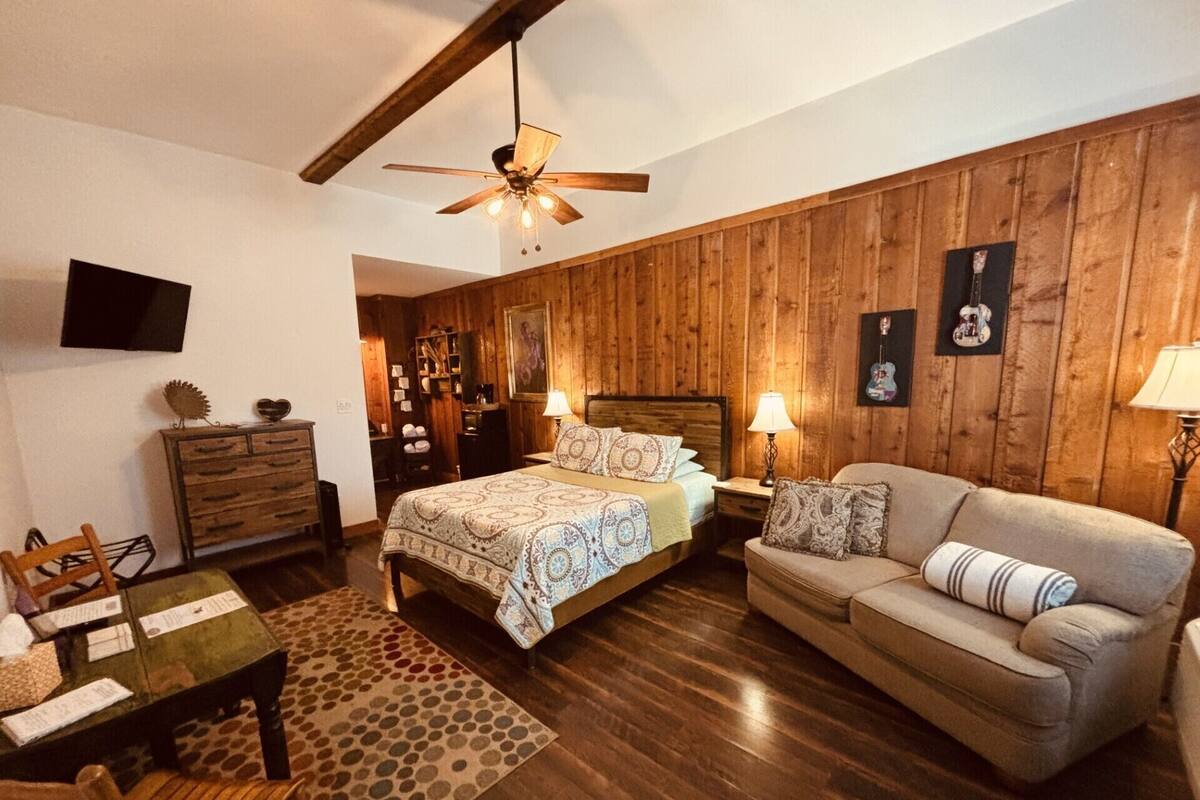 Ein gemütliches Schlafzimmer mit Holzwandverkleidung, einem Bett mit gemusterten Textilien, einem Sofa, einem Schreibtisch mit Stühlen und einem Deckenventilator.