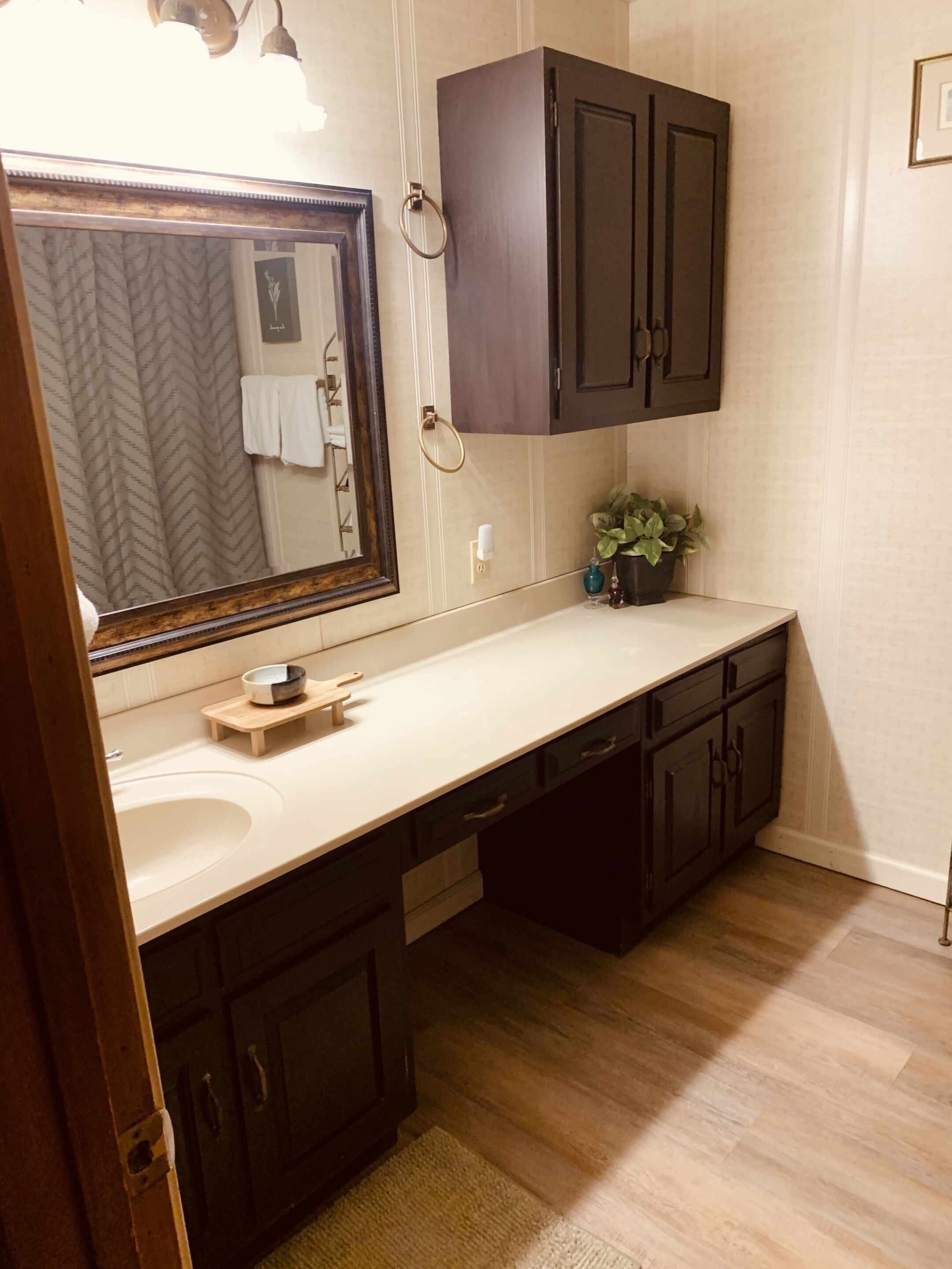 Un baño con un espejo grande, armarios oscuros, una encimera blanca y suelo de madera.